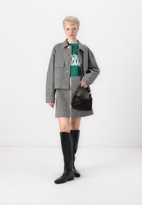 Chaqueta de pana gris con bolsillos parche sobre una camiseta gráfica verde, combinada con una falda de pana a juego y botas negras hasta la rodilla. Bolso marrón.