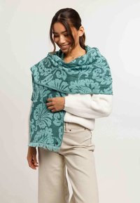 Donna che sorride indossando una sciarpa a tema floreale color teal sopra un maglione bianco e pantaloni beige, su uno sfondo semplice.
