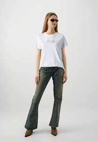 Bela bombažna majica s pisanimi napisi "happy", kombinirana z izpranim razširjenim jeansom in rjavimi škornji s peto.