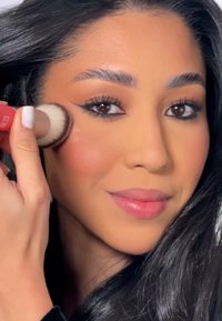 Ein Rouge-Pinsel mit weichen Borsten wird auf die Wange aufgetragen, das Model hat dunkles Haar und ein neutrales Make-up mit braunen Augen und einem hellrosa Lippenstift.