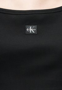 Μαύρο ριμπ ύφασμα με ένα τετράγωνο μαύρο ετικέτα ραμμένη κοντά στη γραμμή λαιμού, που φέρει λευκό λογότυπο "CK Calvin Klein".