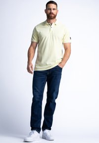 Jasnożółta koszulka polo z kołnierzykiem i dwoma guzikami, w zestawie z ciemnoniebieskimi dżinsami i białymi trampkami.