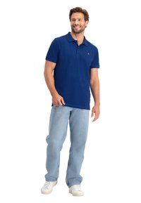 Marineblauwe polo met een kraag, korte mouwen en een groen logo-detail; gedragen met lichtblauwe jeans en witte sneakers.