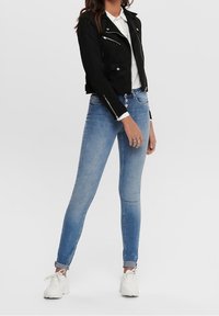Zwarte cropped jas van suède met zilveren ritsen, wit overhemd met kraag, lichtblauwe skinny jeans en witte sneakers.