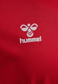 Maglia sportiva rossa Hummel con tessuto texturizzato. Presenta un logo bianco che assomiglia a un'ape e il nome del marchio "hummel" sotto di esso.