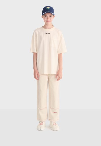 Jeune personne portant un t-shirt oversize crème, un pantalon large crème avec des surpiqûres rouges, des baskets blanches et une casquette bleu marine avec un motif floral.
