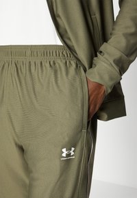 Olivnozeleni trenirki iz gladkega materiala, z elastičnim pasom, stranskimi žepi in belim logotipom Under Armour na levi nogi.