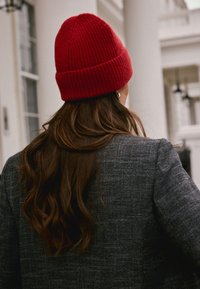 Red knitted beanie with a folded cuff, worn with dark textured blazer. Brown hair cascades down, emphasizing the hat's vibrant color.