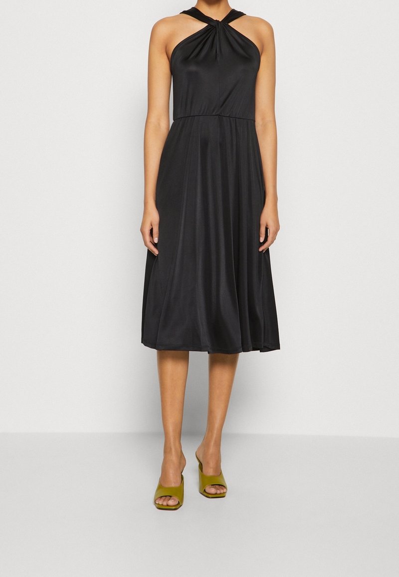 Femme portant une robe midi noire sans manches avec un col halter torsadé et des talons ouverts vert olive, debout devant un fond uni.