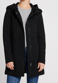 Black, ribbed-textured coat with a hood, front zipper, and snap closure. Worn over a gray top, with pockets and a relaxed silhouette.