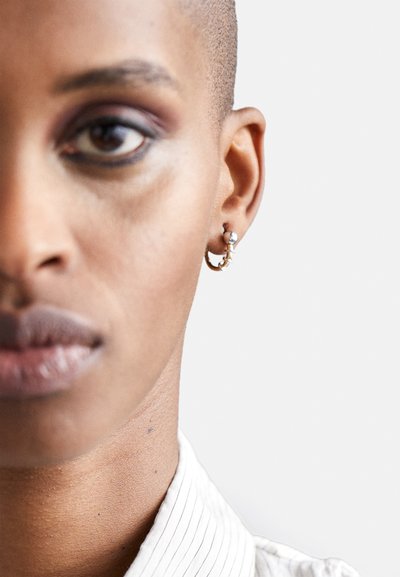 Guld hoopsørering med en sølvperle, der viser et strømlinet design. Baggrunden er neutral og fremhæver øreringens detaljer og bærerens nakke.