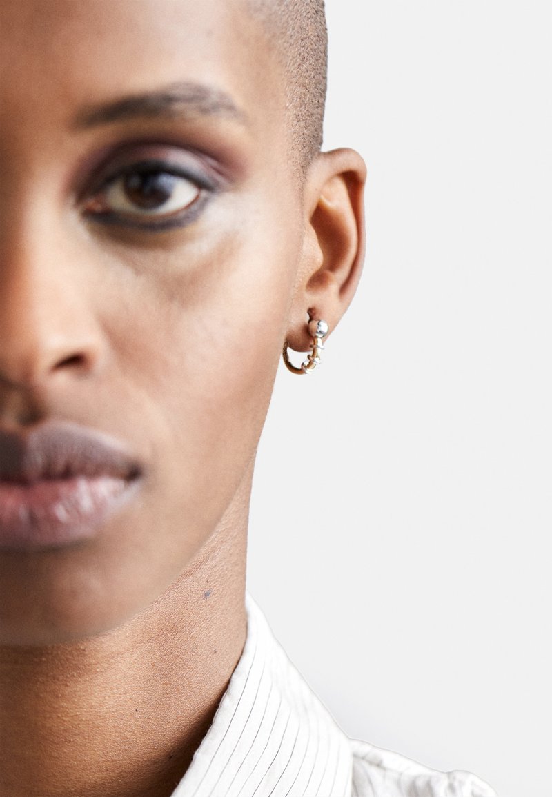 Złota kolczyk w kształcie obręczy z srebrną kulką, prezentująca elegancki design. Tło neutralne, które podkreśla detale kolczyka oraz szyję noszącej.