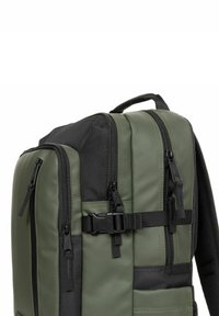 Mochila verde caqui con acentos negros, tela texturizada, múltiples compartimentos con cremallera y correas ajustables. Cuenta con un asa en la parte superior.