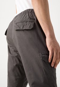 Gros plan d'une personne portant un pantalon cargo gris foncé avec poches à rabat et taille élastique, main reposant sur la cuisse droite.