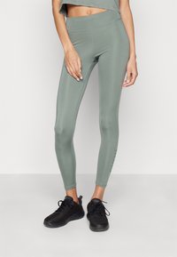 Leggings verdi a vita alta realizzati in tessuto elasticizzato, con una texture liscia e un logo accentato sulla parte inferiore della gamba sinistra, abbinati a sneakers nere.