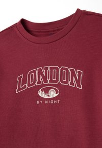 Camiseta de algodón burdeos con cuello redondo. Presenta "LONDRES" en letras blancas negritas y "DE NOCHE" debajo de una ilustración de lugares emblemáticos.