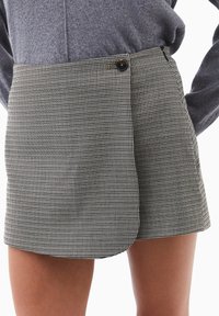 Nærbillede af en person iført en sort-hvid houndstooth omslagsskjørt med en sort knap og en grå sweater.