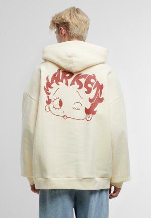 Żółta, oversize bluza z kapturem z dużą czerwoną grafiką kreskówkowej twarzy na plecach, z falującymi włosami i wyolbrzymionymi cechami twarzy.