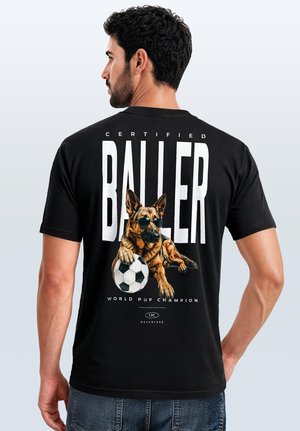 Man with dark hair and beard wearing black t-shirt with graphic of dog in sunglasses holding soccer ball and large text "BALLER" on back.