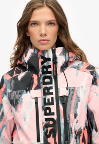 Kolorowa, wzorzysta wodoodporna kurtka z wysokim kołnierzem, zapinanymi kieszeniami i wyraźnym logo "SUPERDRY" w czerni i pomarańczy.