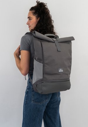 Rucsac gri cu închidere tip roll-top, fabricat dintr-un material durabil, cu un design fluid. Dispune de buzunare laterale și o etichetă cu logo. Bretele ajustabile.