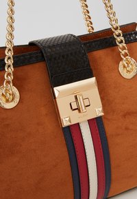 Sac à main en daim marron avec rabat noir texturé, fermoir rectangulaire doré, anses chaîne dorées et rayures verticales rouges, blanches et bleues.