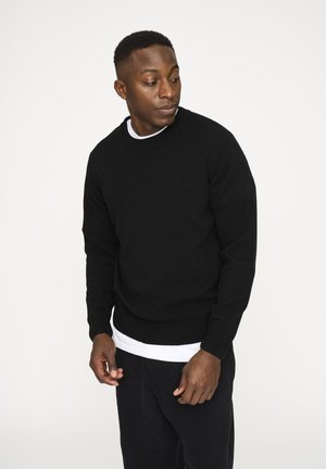 Sort strikket sweater med rund halsudskæring og ribbede manchetter, båret over en hvid skjorte. Stoffet ser blødt og mellemvægtigt ud.
