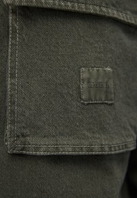 Olivgrüner Denimstoff mit strukturierter Oberfläche. Verfügt über ein quadratisches Patch mit "mavi"-Logo und kontrastierenden Nähten entlang der Kanten.