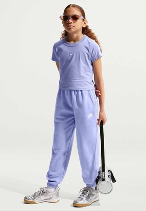 Dziecko ubrane w fioletowy dres Nike, srebrne trampki i okulary przeciwsłoneczne, trzymające małą srebrną cylindryczną torbę na czarnym pasku.