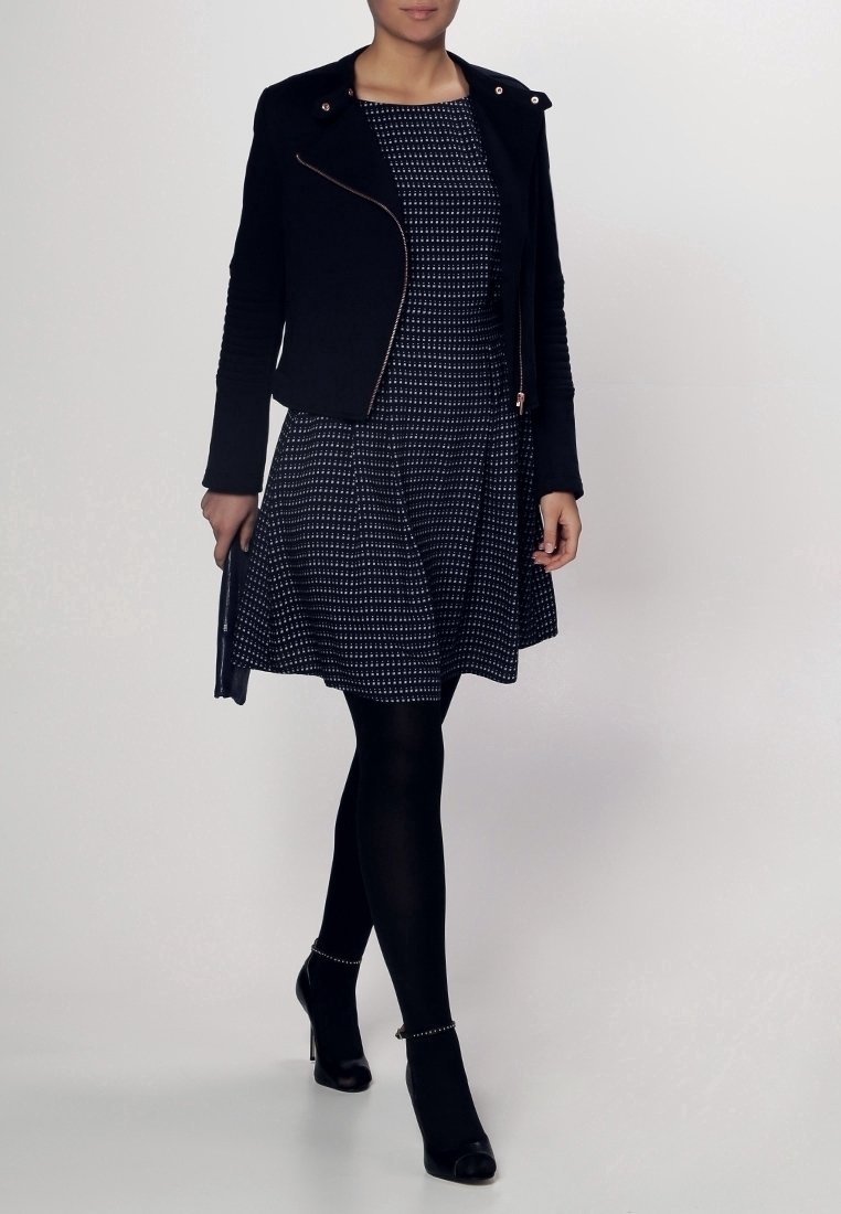 Veste de style motard noire avec des détails zippés en or rose, portée sur une robe navy à pois blancs, associée à des collants noirs et des talons.