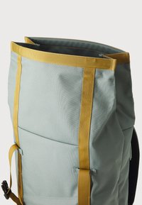 Zaino azzurro chiaro con fibbie gialle, forma strutturata, tessuto resistente e apertura in cima. Presenta tasche esterne e accessori di sicurezza.