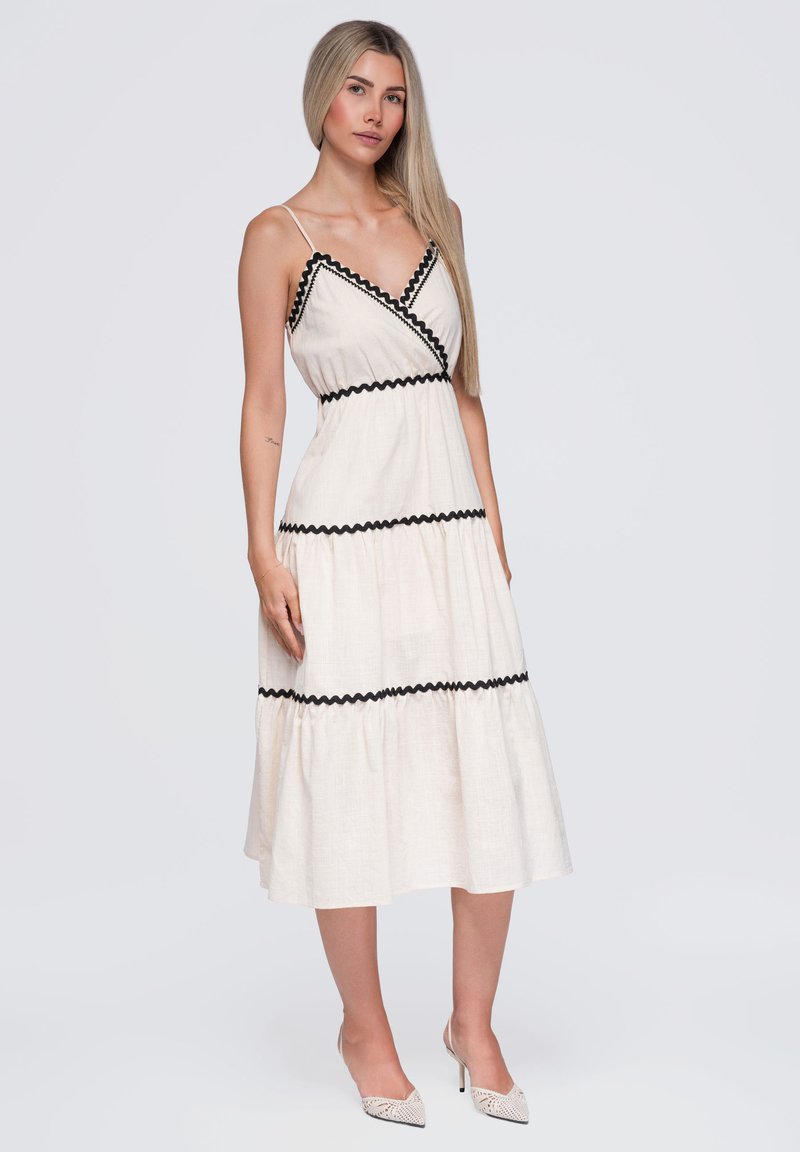 Jeune femme aux longs cheveux blonds portant une robe midi blanche sans manches avec une bordure zigzag noire et des talons blancs pointus sur un fond uni.