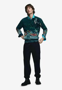 Pletený sveter so zeleným a farebným prírodným vzorom, v kombinácii s čiernymi cargo nohavicami a čiernymi čižmami. Modrá košeľa trčí z goliera svetra.