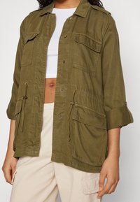 Osoba ubrana w oliwkowo zieloną kurtkę roboczą z patkami kieszeni i ściągaczami, na białym krótkim topie oraz beżowych spodniach typu cargo.