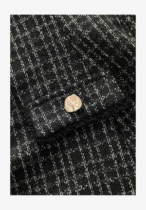 Zwart-wit gestructureerde stof met een ruitpatroon, voorzien van een gouden metalen knoopaccent aan de voorkant. Zacht, geweven materiaal.