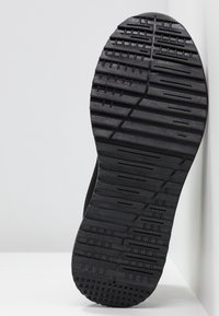 Semelle de chaussure en caoutchouc noir avec des motifs géométriques variés conçus pour l'adhérence, positionnée debout contre un fond blanc et un coin de mur.