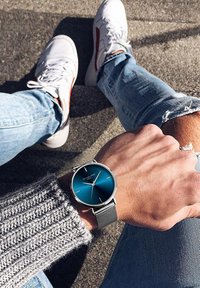Orologio da polso argento con quadrante blu e cinturino in maglia grigia, indossato su una mano accanto a un maglione grigio con texture e jeans blu. Sneakers bianche visibili.