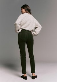 Pantaloni din corduroy verde măsliniu cu talie înaltă, cu buzunare la spate, purtați cu un pulover deschis și pufoș. Văzuți din spate.
