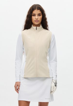 Gilet beige senza maniche con cerniera frontale, abbinato a maniche lunghe bianche. Tessuto liscio con una leggera lucentezza. Gonna bianca con logo sull'orlo.