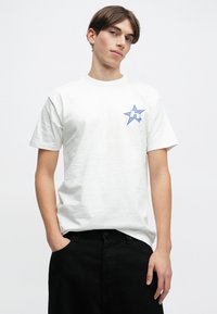 Λευκό βαμβακερό μπλουζάκι με μπλε γραφικό αστέρα και λογότυπο 'h' στο στήθος. Κοντά μανίκια, στρογγυλή λαιμόκοψη, άνετη εφαρμογή, συνδυασμένο με μαύρο παντελόνι.