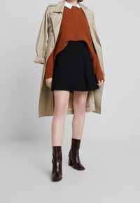 Trench coat beige, suéter de punto color óxido con textura elevada, falda negra acampanada y botines de tacón marrón brillante.