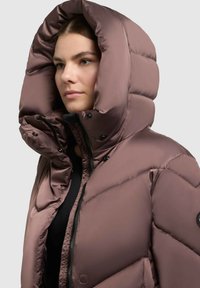 Tupsuline, lavendlililla värvi jope suure kapuutsiga, millel on tõmblukk ja trukid, valmistatud läikivast, tikandiga materjalist.