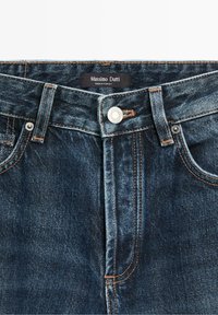 Jean en denim bleu foncé avec fermeture par bouton argenté, passants de ceinture et étiquette indiquant Massimo Dutti fabriqué en Turquie.