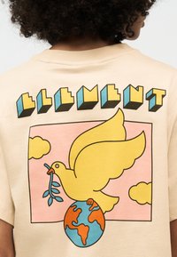 Άτομο που φοράει μπεζ πουκάμισο με γραφικό κίτρινο περιστέρι που κρατά κλαδί ελιάς, που στέκεται πάνω σε γη μπλε και πορτοκαλί, με φόντο ροζ ουρανό.