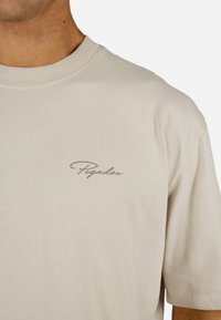 Vaaleanbeige puuvillapaita, jossa on pyöreä kaula-aukko, lyhyet hihat ja huomaamaton, tyylitelty "Pegador"-logo oliivinvihreänä rinnassa.