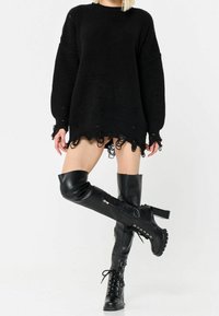 Femme portant un pull noir oversize usé et des bottes noires montantes à lacets avec des talons épais, debout, jambes croisées.