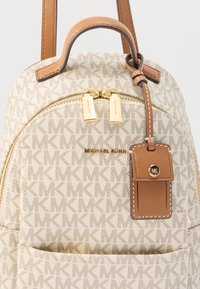Sac à dos beige Michael Kors avec motif monogramme, tirettes de fermeture éclair dorées, poignée en cuir marron, et étiquette en cuir marron attachée avec bouton logo MK.
