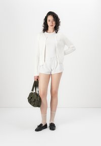 Hvit kabelstrikket cardigan over en tettsittende hvit topp, parret med lyse striper shorts og svarte sko. Holder en grønn stoffhåndveske.