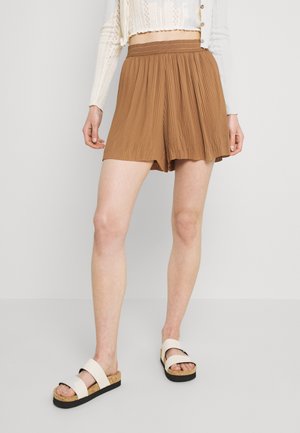 Bruine geplooide korte broek met een elastische tailleband, gemaakt van lichtgewicht stof. Gecombineerd met beige plateauzandalen met twee brede banden.