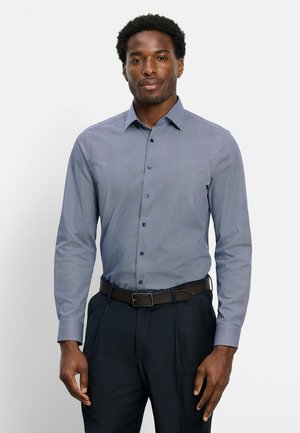 Uomo che indossa una camicia azzurra con motivi a maniche lunghe, infilata in pantaloni scuri con cintura nera, in piedi davanti a uno sfondo bianco.